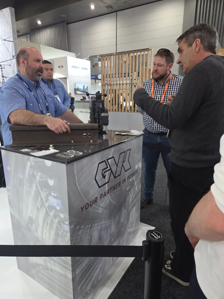 Rail track drilling equipment demonstrated on a length of rail at the G.V. Kinsman stand, with four attendees in conversation.